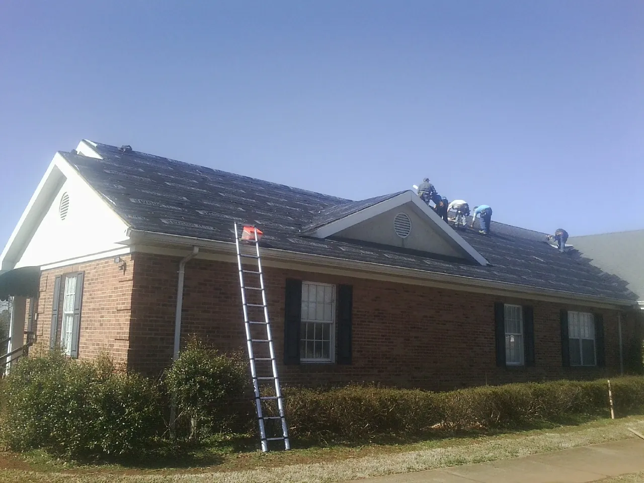 Crew tearing shingles off a brick ranch roof with a ladder set — Emergency Roof Repair work by our crew in Plevna