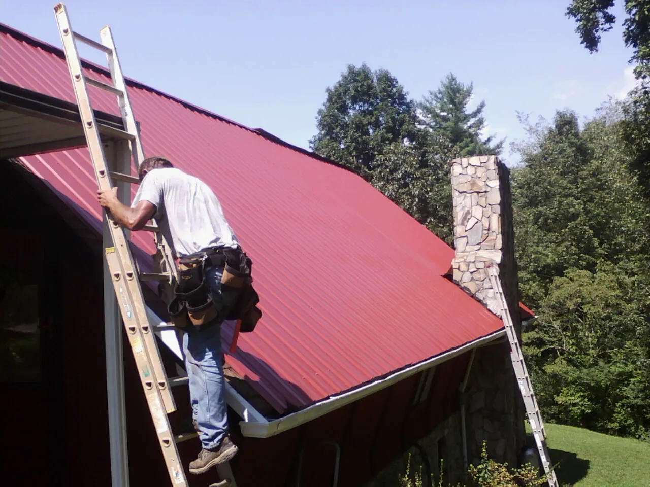 Roofer on a ladder beside a red metal roof and stone chimney — Soffit & Fascia Repair by Roofing Brothers in Plevna
